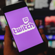 A hand holding a phone with the Twitch logo in front of a screen displaying multiple twitch channels.